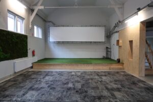 Kleine presentatieruimte in het Gedempt Hamerkanaal met een podium van kunstgras, een groot projectiescherm en een industriële inrichting.